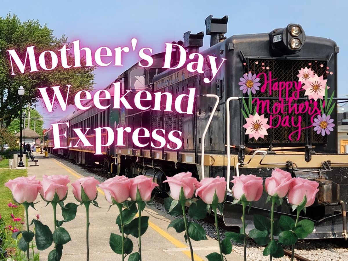 Train with 'Happy Mother's Day' sign, pink roses in front, buildings and blue sky in background.